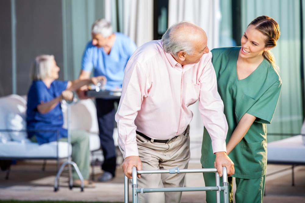 Happy Caretaker Assisting Senior Man In Using Zimmer Frame