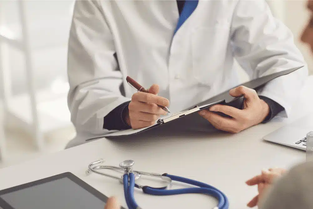 Doctor in white coat writing notes on clipboard with stethoscope on desk