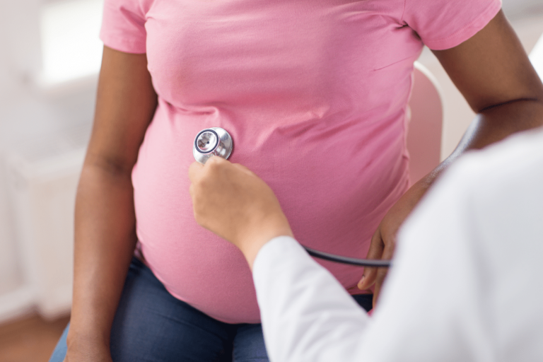 Pregnant woman in a pink shirt visiting a doctor. The doctor has a stethoscope ip to the woman's belly.