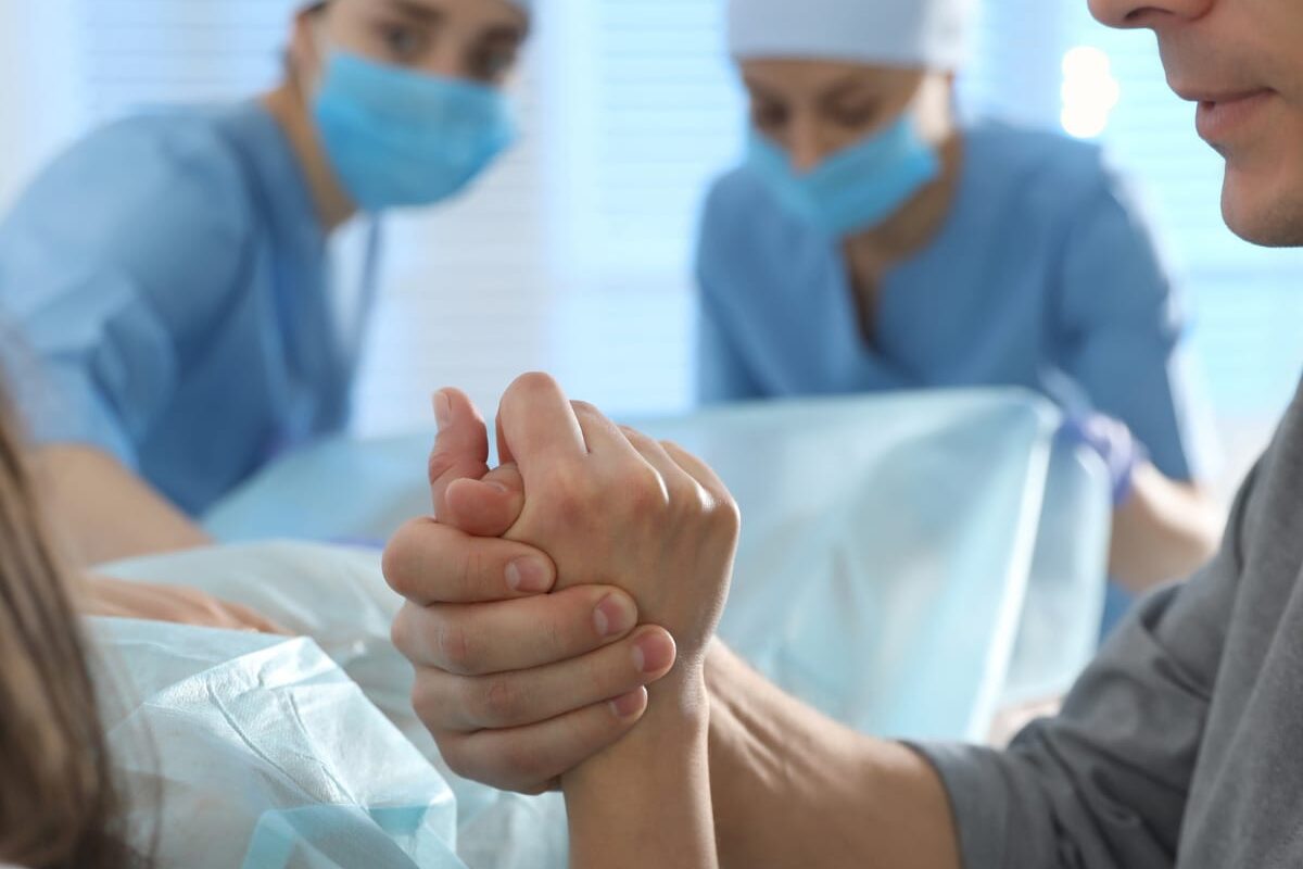 Husband holding wife's hand during labor, with two doctors in the background at the hospital.