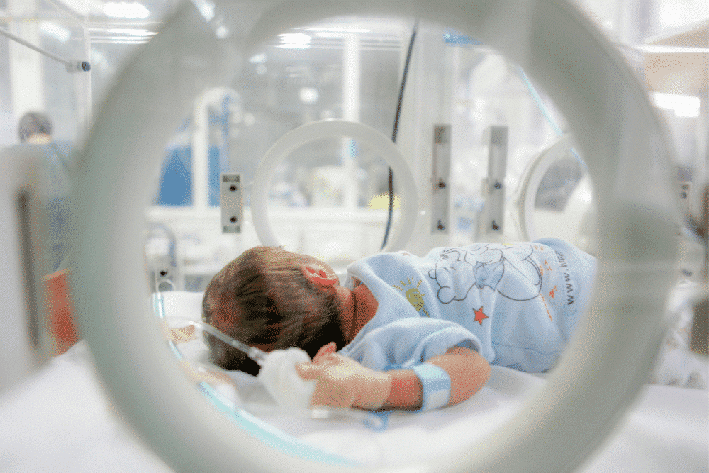 newborn wearing a blue sleeper and a wristband, lying on their side inside a hospital incubator