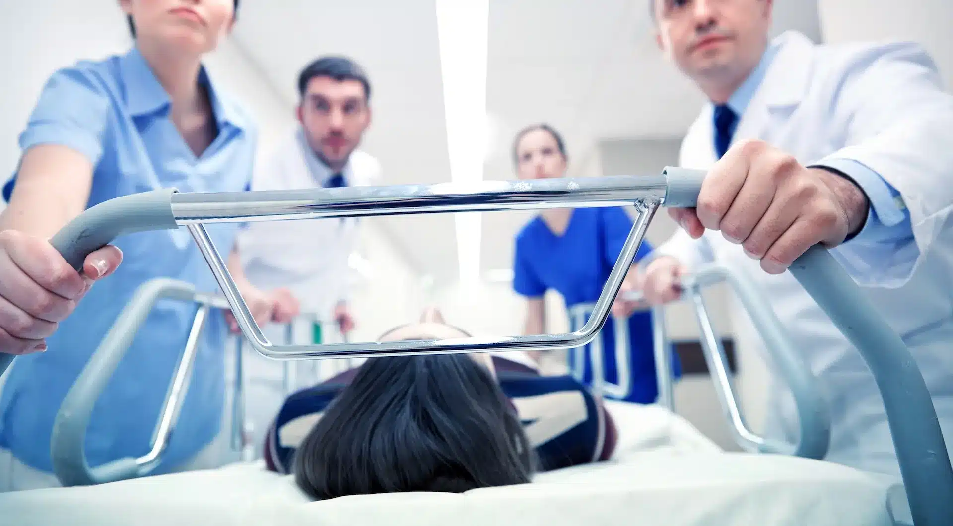 Medical staff rush a patient on a hospital stretcher down a hallway, with the patient lying on their back and doctors and nurses visible in the background.