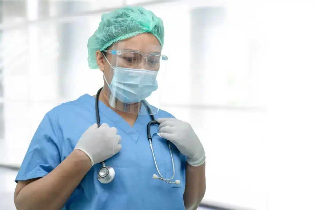 Nurse in blue scrubs wearing surgical mask, face shield, and hair cap with stethoscope in PPE