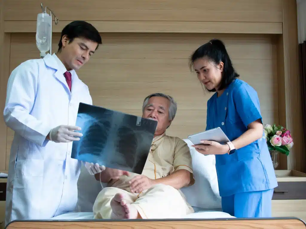 Doctor and nurse reviewing chest X-ray with elderly male patient in hospital bed with IV drip