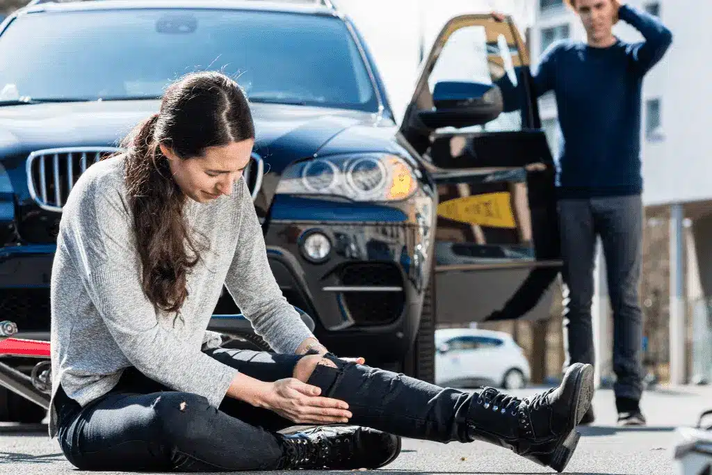 Injured woman sitting on pavement holding her leg after car accident with damaged vehicle