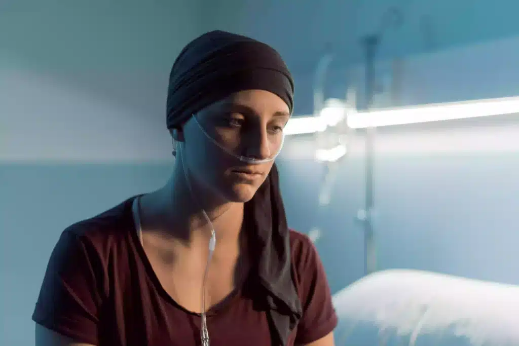Female cancer patient wearing a black headscarf and nasal oxygen tube, sitting in a hospital bed