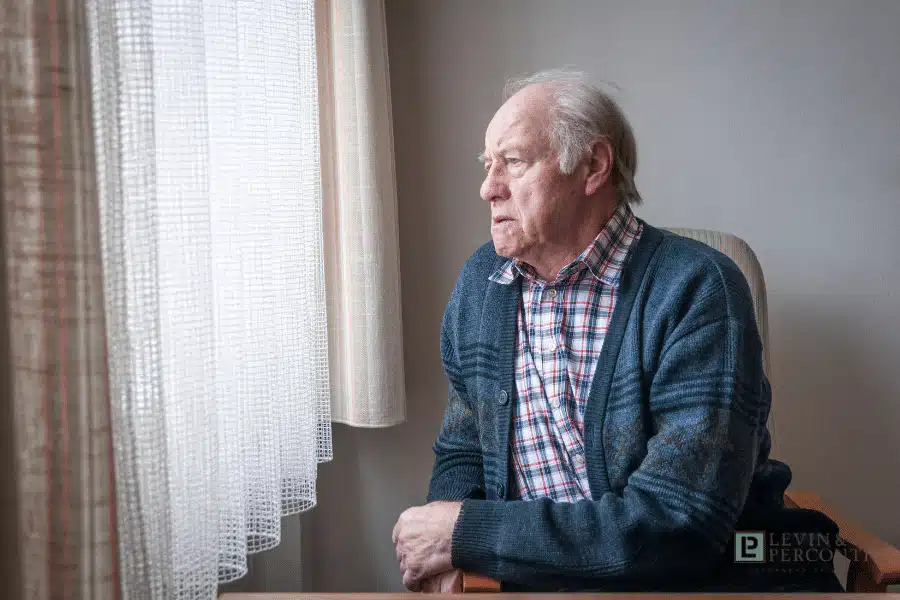 Elderly man in plaid shirt and cardigan sitting pensively by window in residential care setting
