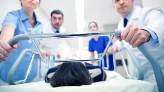 medics-and-patient-on-hospital-gurney-at-emergency Medical staff rush a patient on a hospital stretcher down a hallway, with the patient lying on their back and doctors and nurses visible in the background.